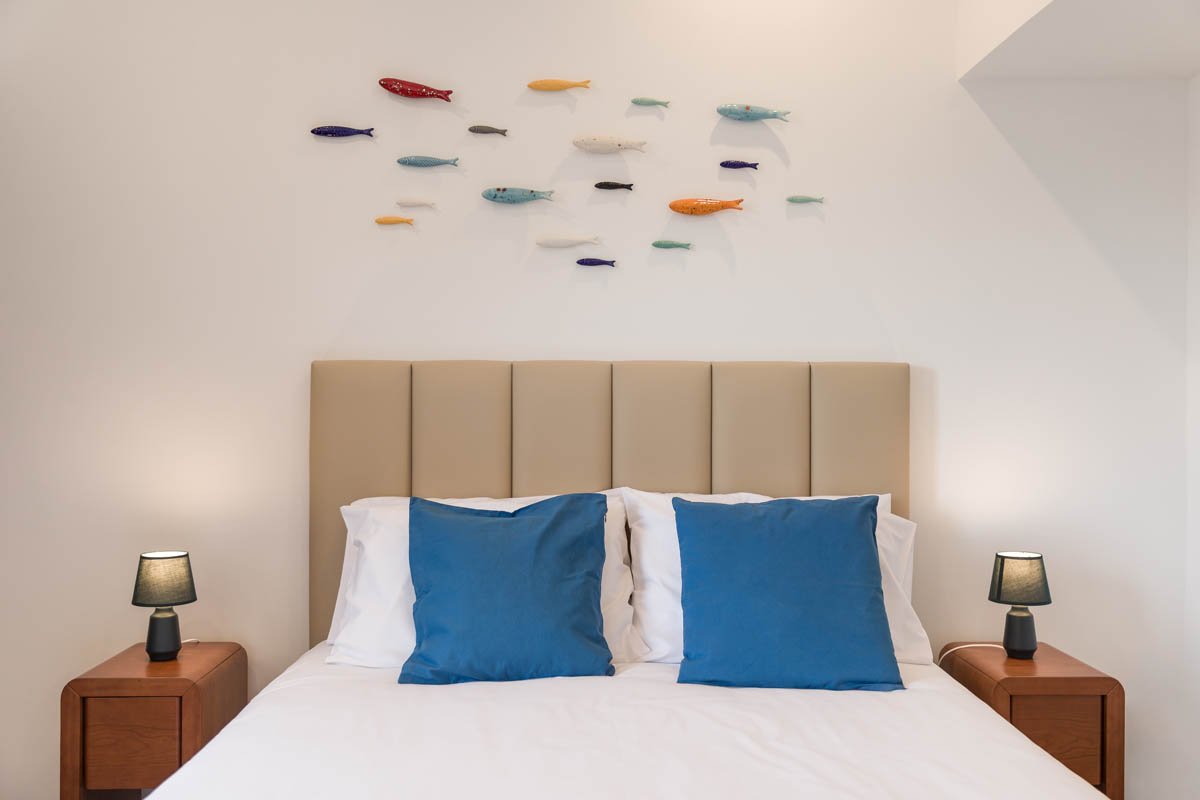 Cama de casal com cabeceira padded bege, duas almofadas azuis, luminárias ao lado em mesas de madeira, parede branca com arte de peixes coloridos.