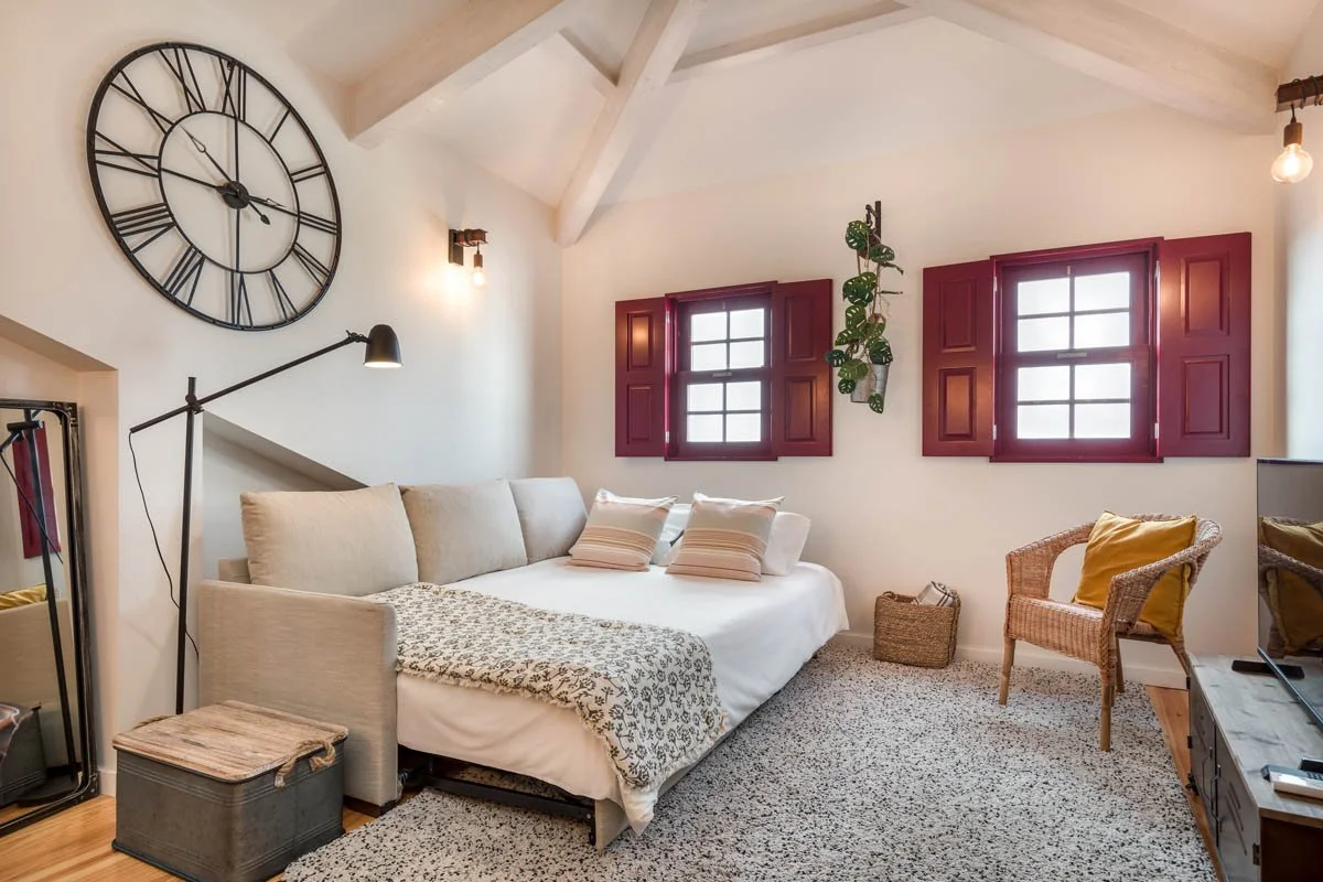 Quarto decorado com paredes brancas, uma janela com persianas vermelhas, um relógio grande de parede, uma cama com travesseiros e um cobertor, uma cadeira de vime com travesseiro amarelo, uma planta pendurada na parede.