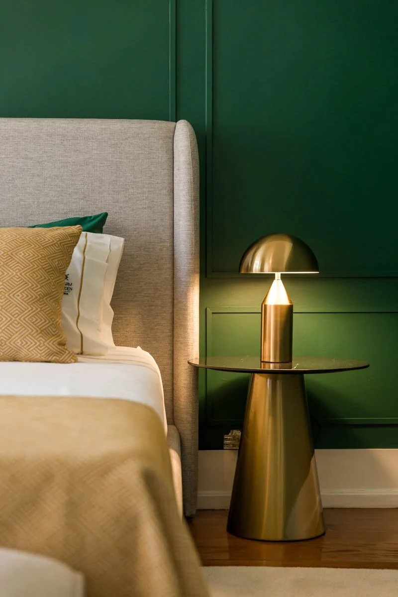 Foto de um quarto com cabeceira de cama acolchoada, almofadas decorativas em tons de dourado e verde, parede verde escura ao fundo, e uma luz de mesa dourada sobre uma mesa de canto também dourada.
