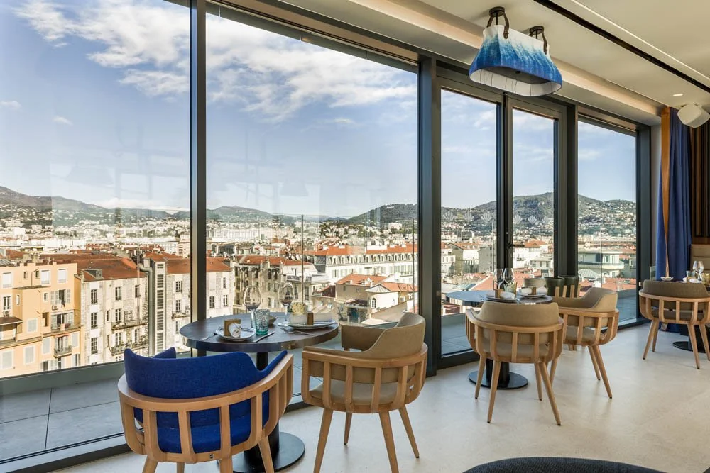 Interior de um restaurante com vista para uma cidade, janelas de vidro grande, mesas com cadeiras de madeira e decoração moderna