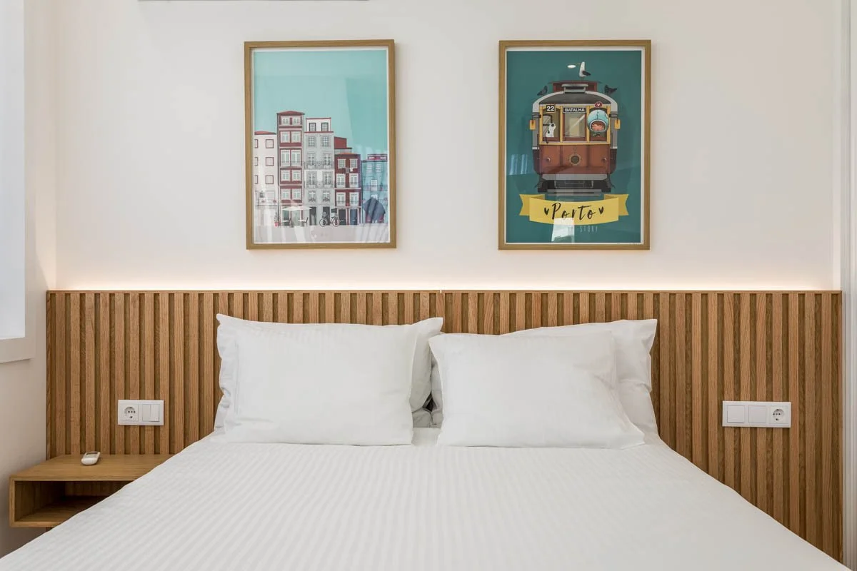 Quarto com cama de casal com cabeceira de madeira, duas almofadas brancas, duas tomadas e um controle remoto na mesa ao lado. Acima da cama, duas molduras com ilustrações de cidades e transporte urbano, uma delas com um bonde e a palavra "Porto".