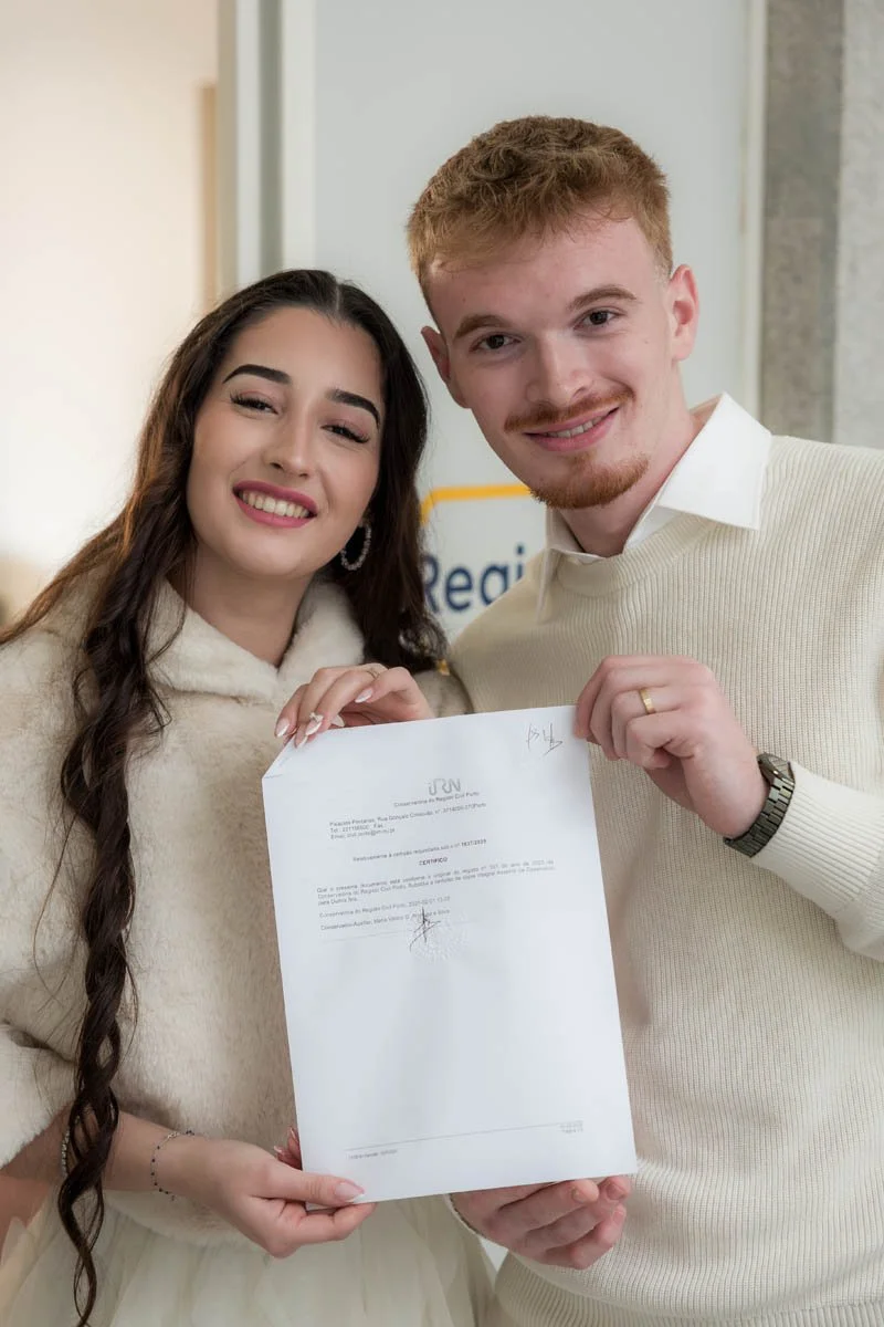 Um casal sorridente segurando um documento ou certificado, em um ambiente interno, possivelmente uma cerimônia ou evento oficial.
