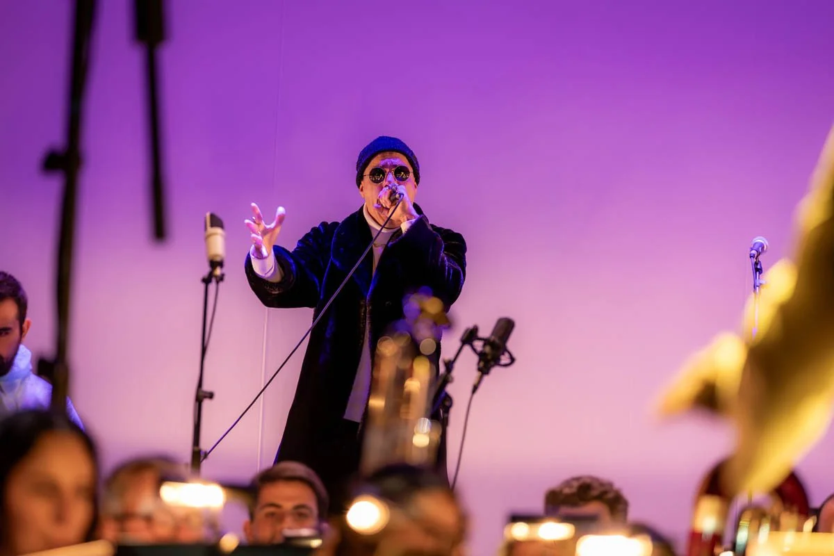 Um cantor usando chapéu, óculos escuros e casaco preto, segurando um microfone durante uma apresentação, com várias pessoas ao redor e microfones no palco.