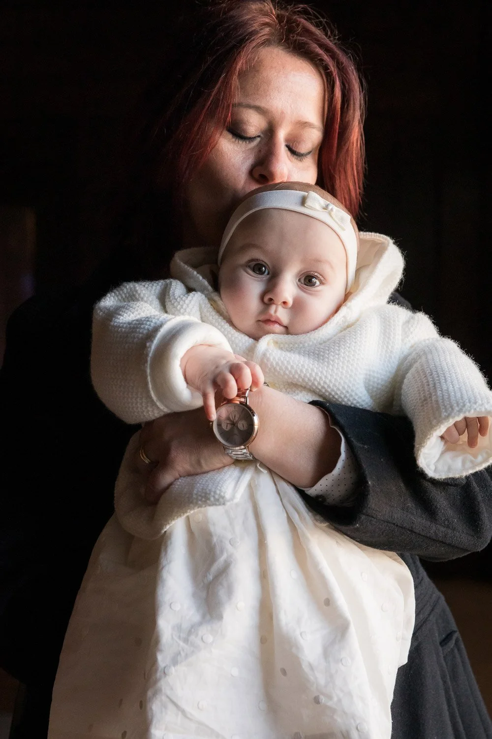 Mulher com cabelo vermelho segurando um bebê com roupas brancas e chapéu, em ambiente interno com luz baixa.