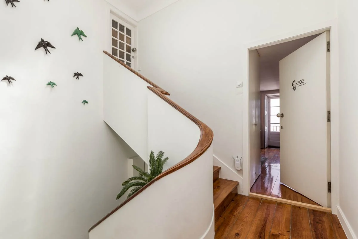 Entrada de um apartamento com paredes brancas, porta aberta à direita, escada de madeira com corrimão curvo e decoração de pássaros de metal na parede esquerda.