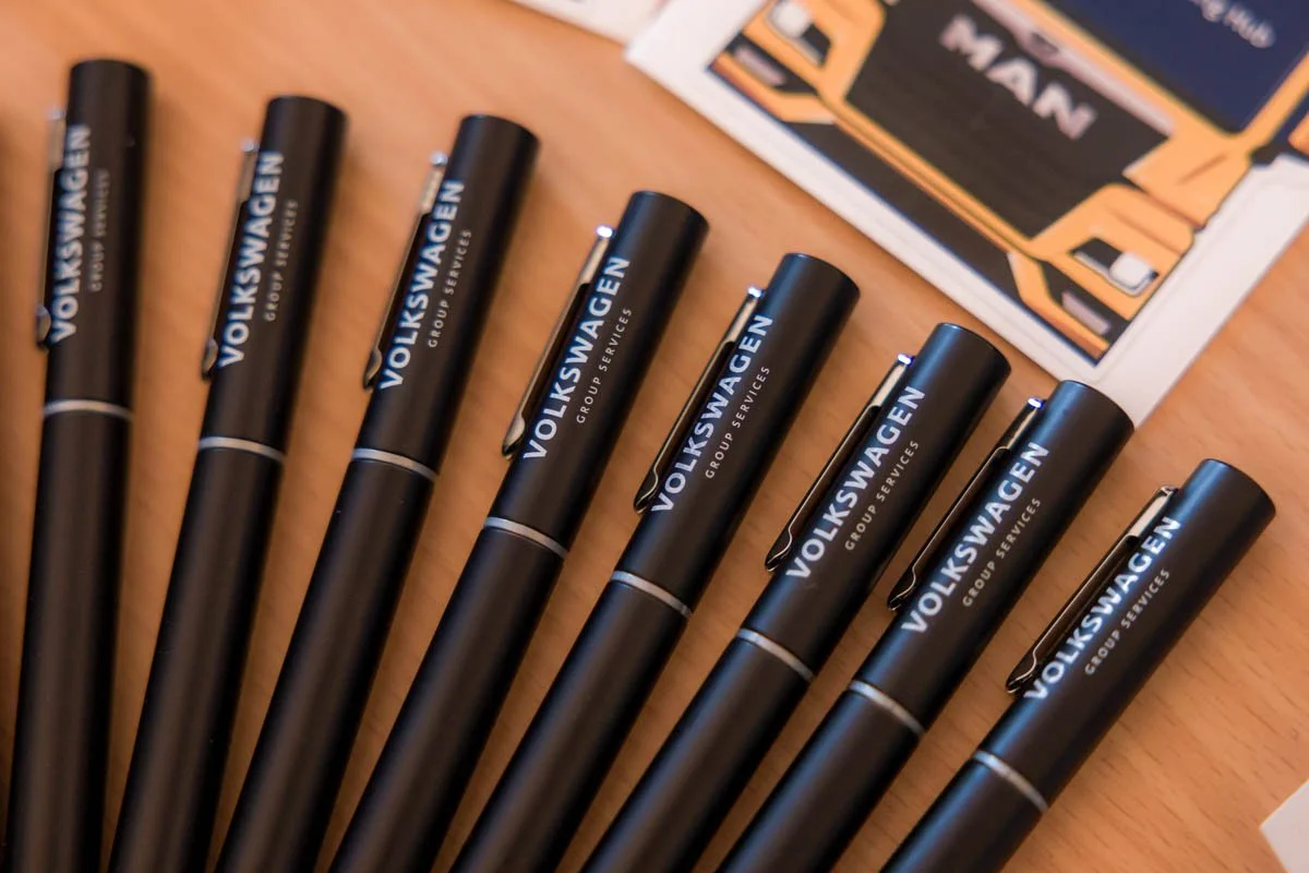 Grupo de marcadores pretos com a marca Volkswagen em destaque, alinhados em uma mesa de madeira, com uma caixa de cores na parte superior direita.