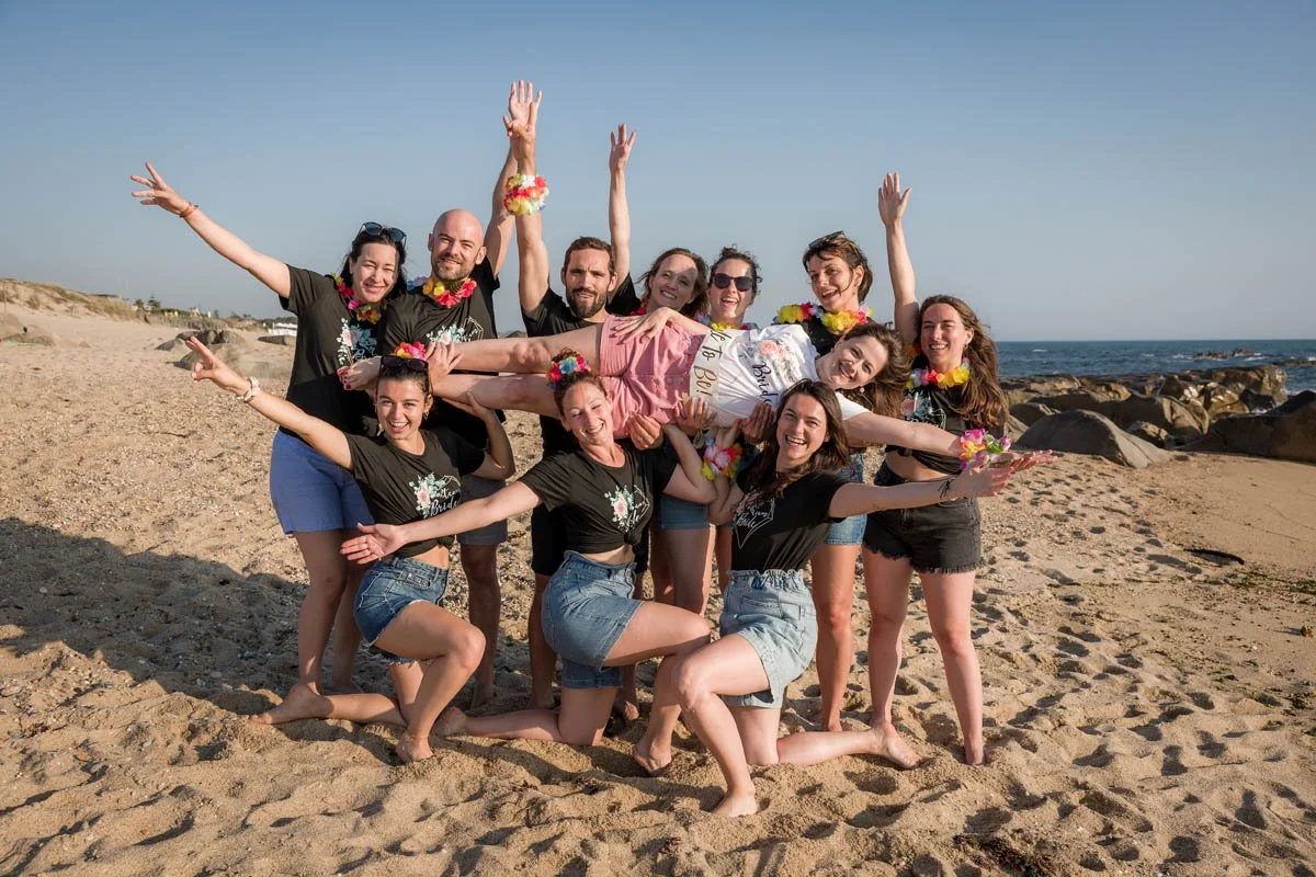 Grupo de amigos na praia, celebrando, com uma mulher sendo carregada por outros na comemoração de despedida de solteira, usando colares de flores.
