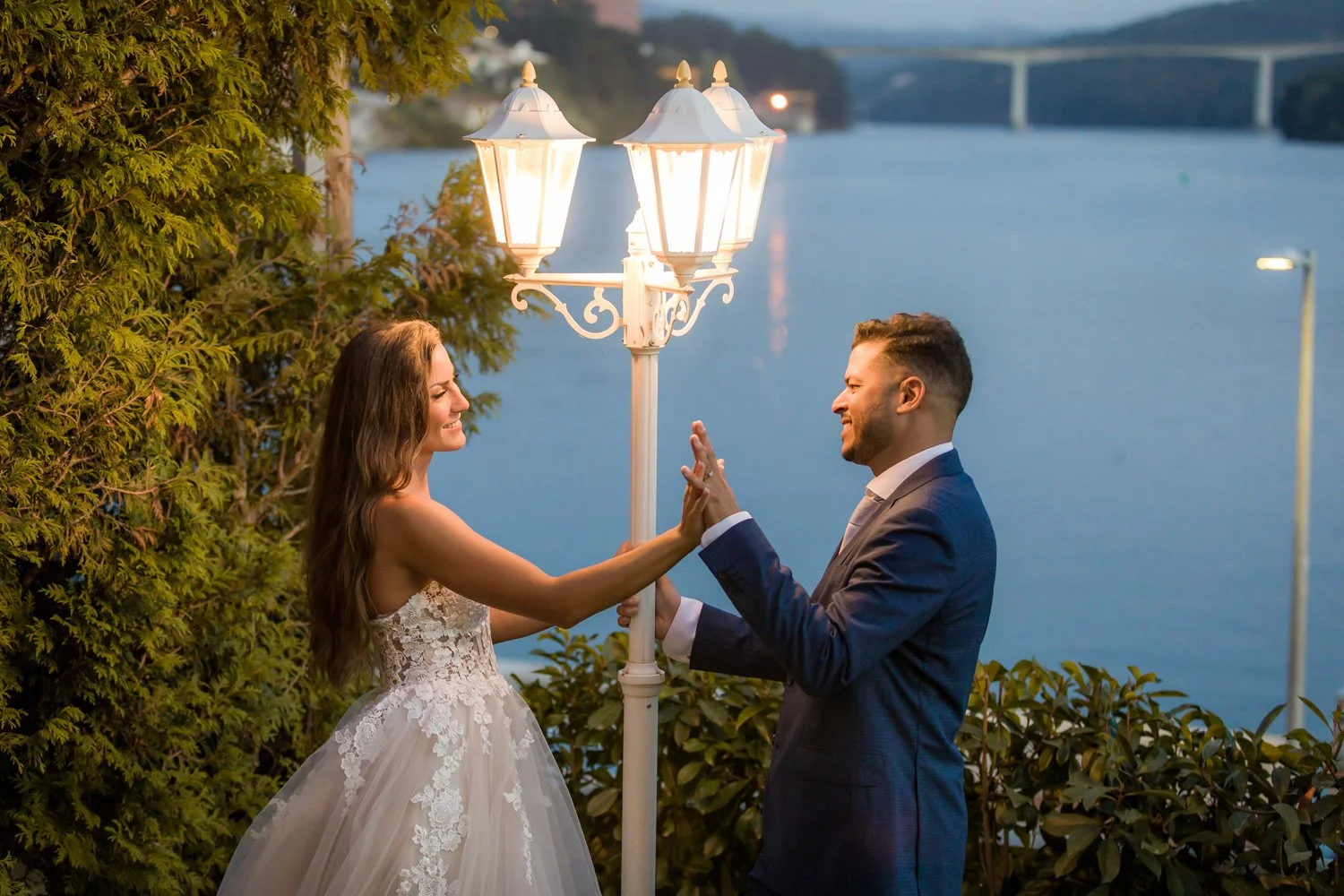 Casal de casamento ao pôr do sol, com a noiva vestindo vestido de casamento branco e o noivo de terno, segurando uma luminária antiga entre eles, sorrindo e tocando as mãos na frente de uma paisagem aquática com uma ponte ao fundo.