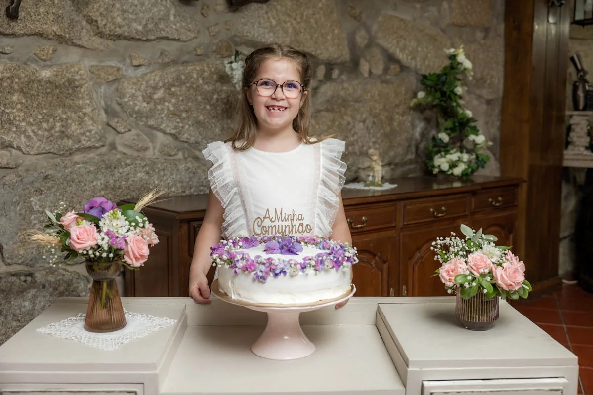 Menina de óculos segurando um bolo de aniversário decorado com flores, com um topo escrito "Minha Primeira Comunhão", ao lado de vasos com flores em uma mesa de jantar de madeira, ao fundo parede de pedra.