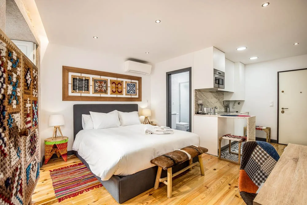 Quarto de hotel com cama confortável, decoração moderna, paredes brancas, piso de madeira, pequena cozinha ao lado com utensílios, iluminação embutida e detalhes decorativos coloridos.