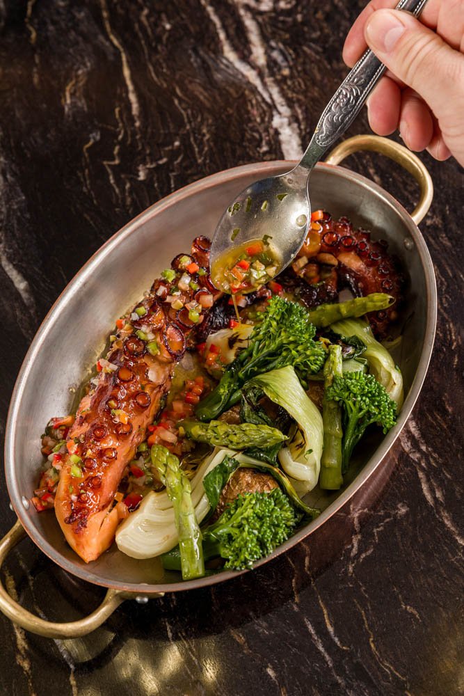 Peixe com molho de ervas, acompanhado de legumes cozidos, em uma travessa de metal com alças douradas