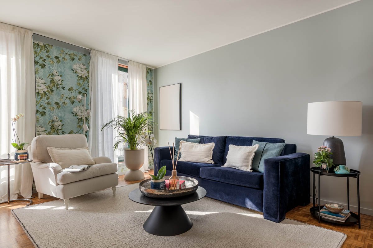 Sala de estar com sofá azul, poltrona branca, mesa de centro preta, luminária com base escura, plantas, janelas com cortinas brancas e papel de parede floral.