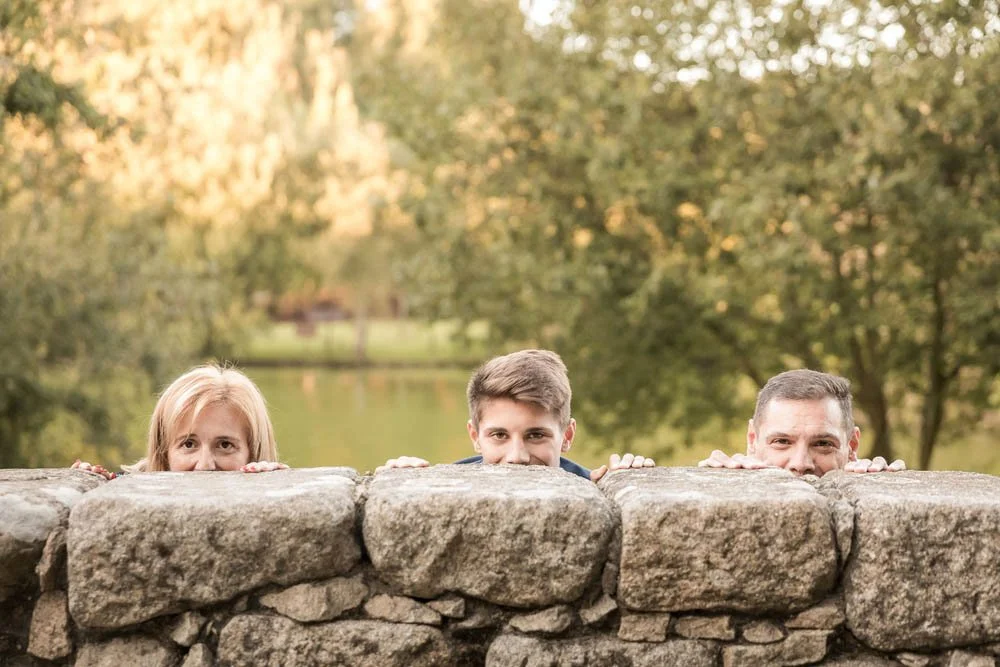 Três pessoas, um idoso, um jovem e um adulto, escondendo-se atrás de uma parede de pedra em um parque com árvores ao fundo.