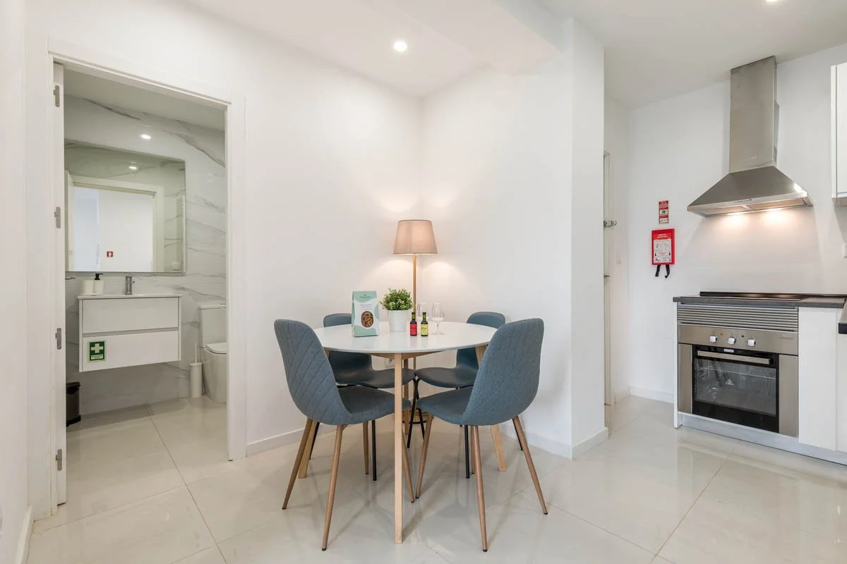 Sala de jantar com mesa redonda, quatro cadeiras azuis, luminária de chão com abajur, cozinha com fogão e exaustor, porta para banheiro ao lado, parede branca.