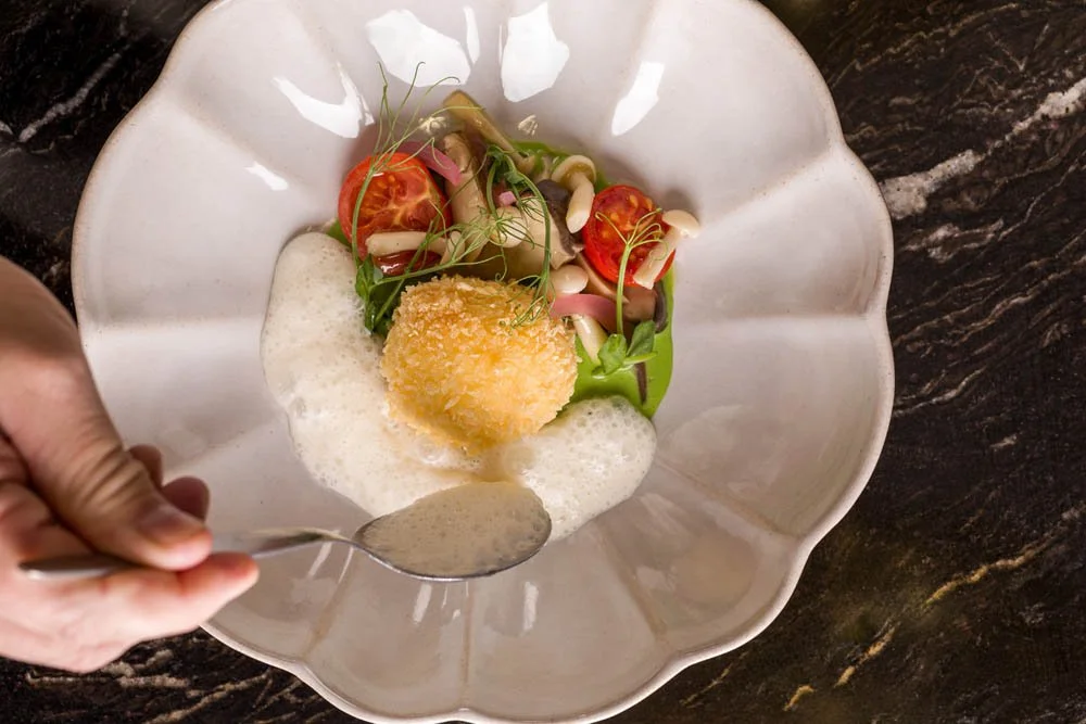 Prato de comida gourmet com uma croqueta dourada, tomates, folhas verdes, brotos, e um molho branco, servido em uma tigela branca com um fundo de mármore escuro.