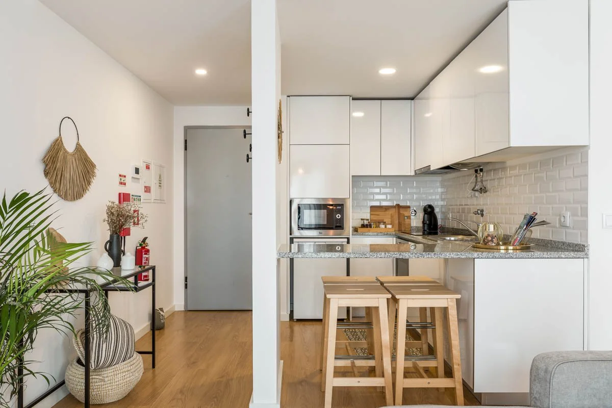 Cozinha moderna com bancada de granito, armários brancos e parede de azulejos brancos. Dois taburetes de madeira na frente da bancada, eletrodomésticos visíveis, incluindo micro-ondas e cafeteira. Decor de parede com objetos de decoração e plantas ao