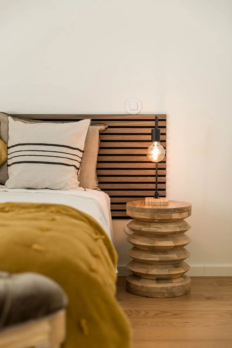 Quarto com cabeceira de madeira, travesseiros com capas de listras, manta amarela e uma luminária de abajur de vidro no criado-mudo de madeira, com parede branca ao fundo.