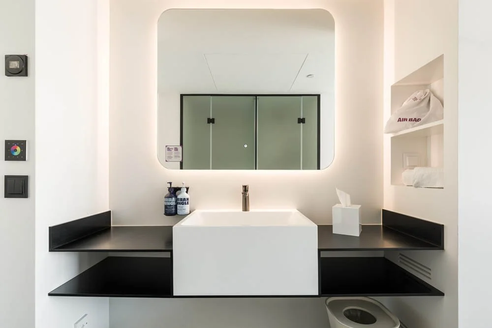 Banheiro com espelho grande, pia branca, torneira moderna, prateleiras pretas, sabonetes, papel toalha, sacola de aeroporto no nicho, sabonete líquido, toalhas e uma peça de roupa na prateleira.