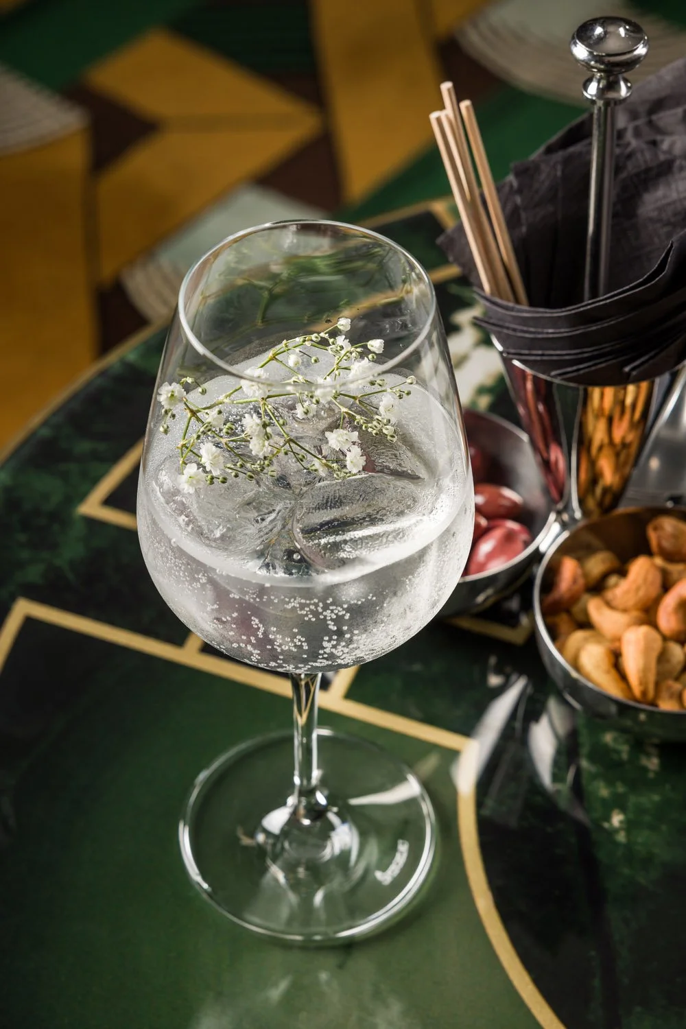 Copo de drink transparente com gelo e florzinha branca decorativa, ao lado de uma bandeja com amendoins, guardanapos, canudos de bambu, e canudo de metal, tudo colocado sobre uma mesa de cor verde escura.