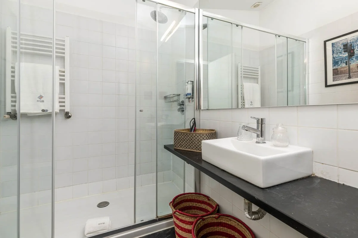 Banheiro com box de vidro, pia retangular branca, espelho grande, toalhas brancas, uma cesta de artesanato com itens ao lado da pia, pisos de azulejos brancos, um quadro na parede e cestos de vime na frente da pia.