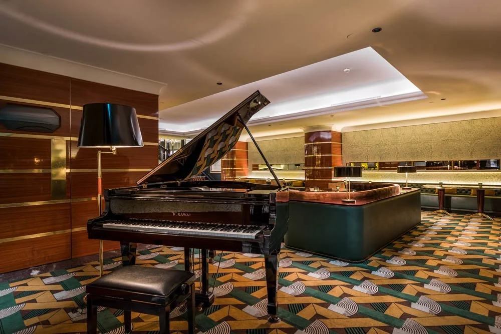 Salão com piano de cauda preto, luminária de chão preta com iluminação suave, parede de madeira e parede com iluminação embutida, piso com tapete de padrão geométrico