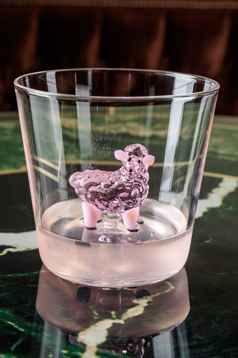 Vaso transparente com uma peça de decoração em forma de carneiro feita de vidro rosa e detalhes em branco, apoiado em uma mesa de tampo de mármore escuro.