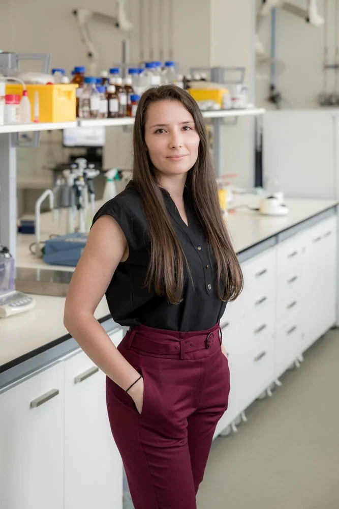 Mulher jovem com cabelo castanho, vestindo blusa preta e calças vermelhas, posando em laboratório com bancadas, equipamentos e frascos de vidro ao fundo.