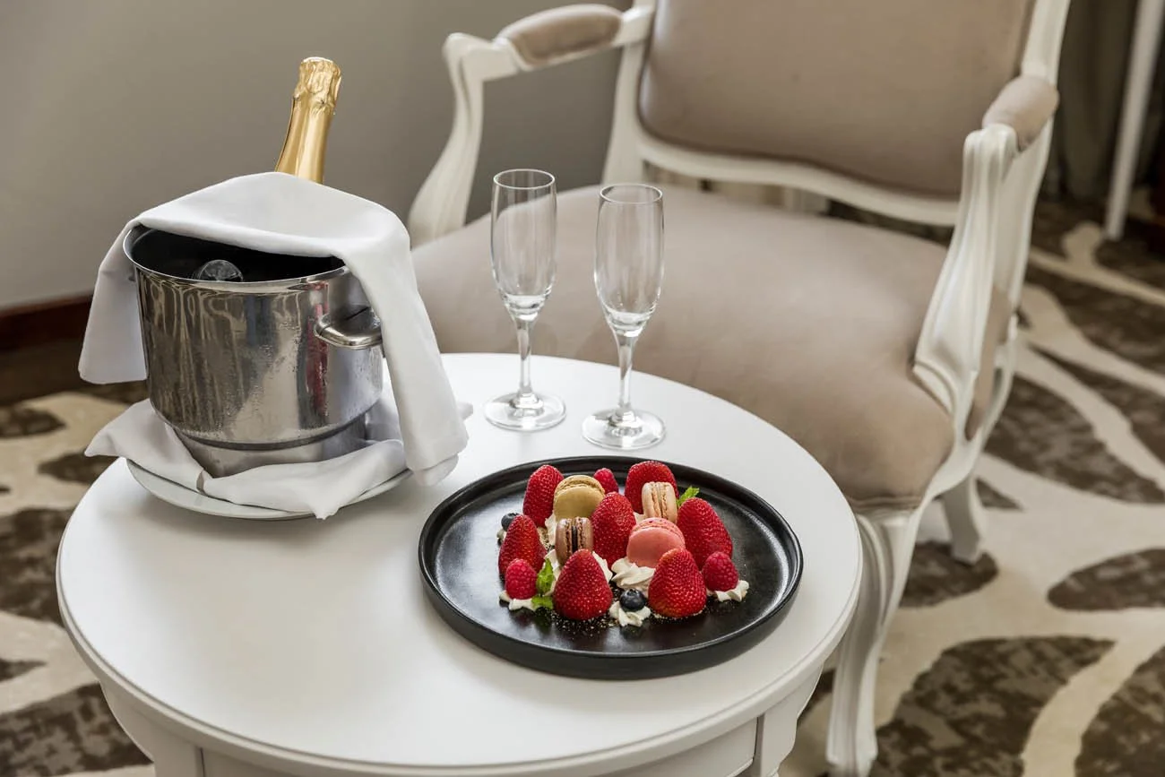 Mesa de hotel com champanheira, duas taças de champanhe, ouros e um prato com diferentes tipos de sobremesas, incluindo morangos, macarons e frutas.