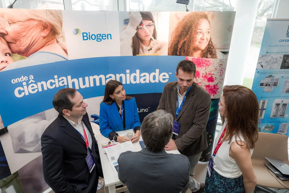 Grupo de cinco pessoas conversando na feira de ciências da Biogen, com banner ao fundo dizendo 'onde a humanidade'.