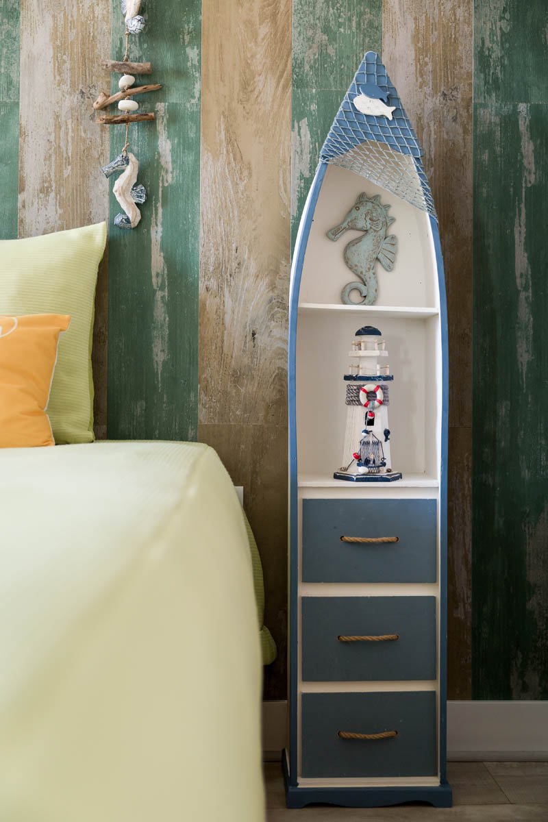 Cabideiro decorativo em forma de barco com enfeites de decoração náutica e um berço ao lado com travesseiros de cor amarela e laranja. Fundo de parede com pintura de efeito envelhecido em cores verdes e bege.