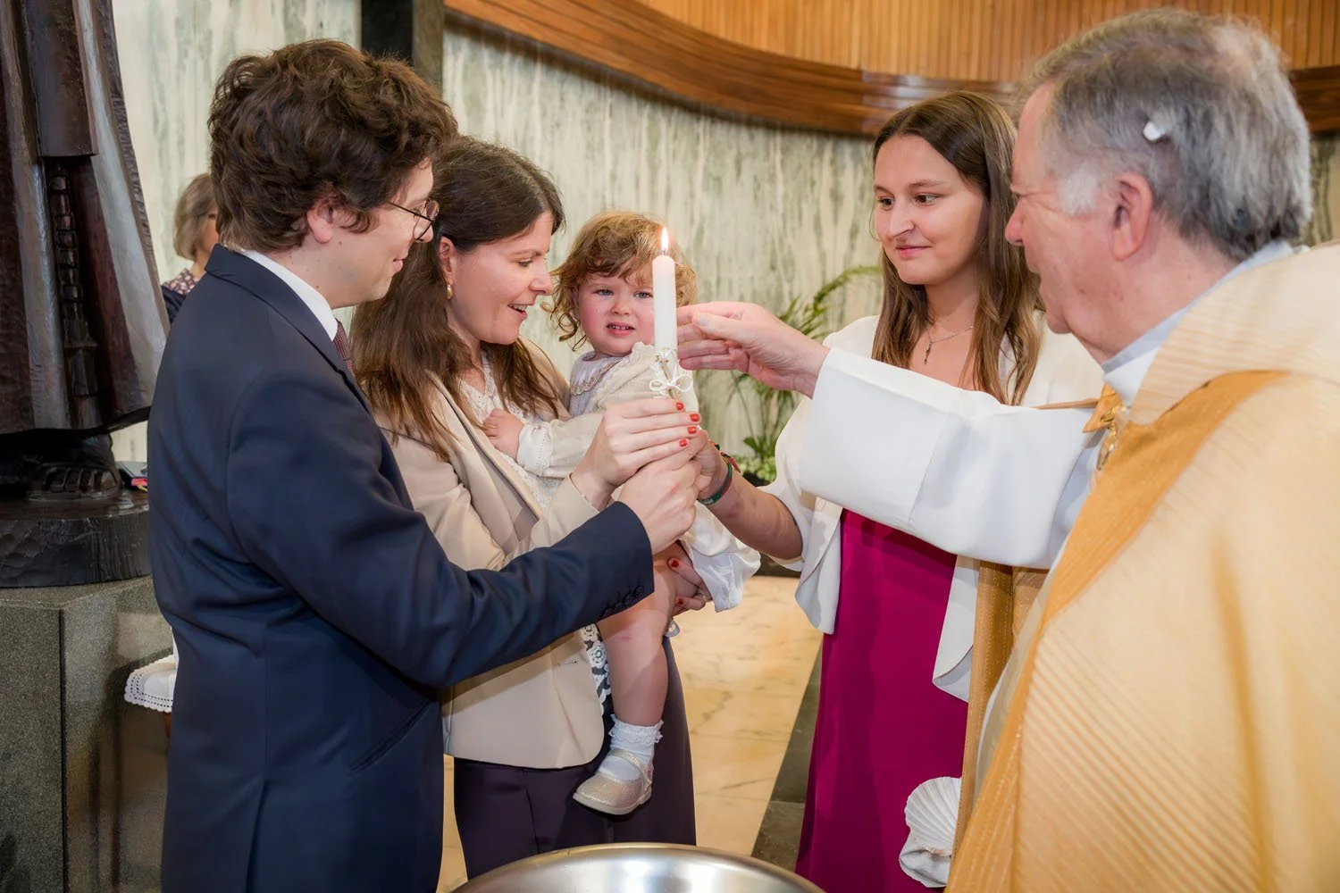 Cerimônia religiosa de batismo com um padre, uma mãe segurando uma criança com uma vela e uma madrinha ao lado, enquanto um jovem ajuda na cerimônia.