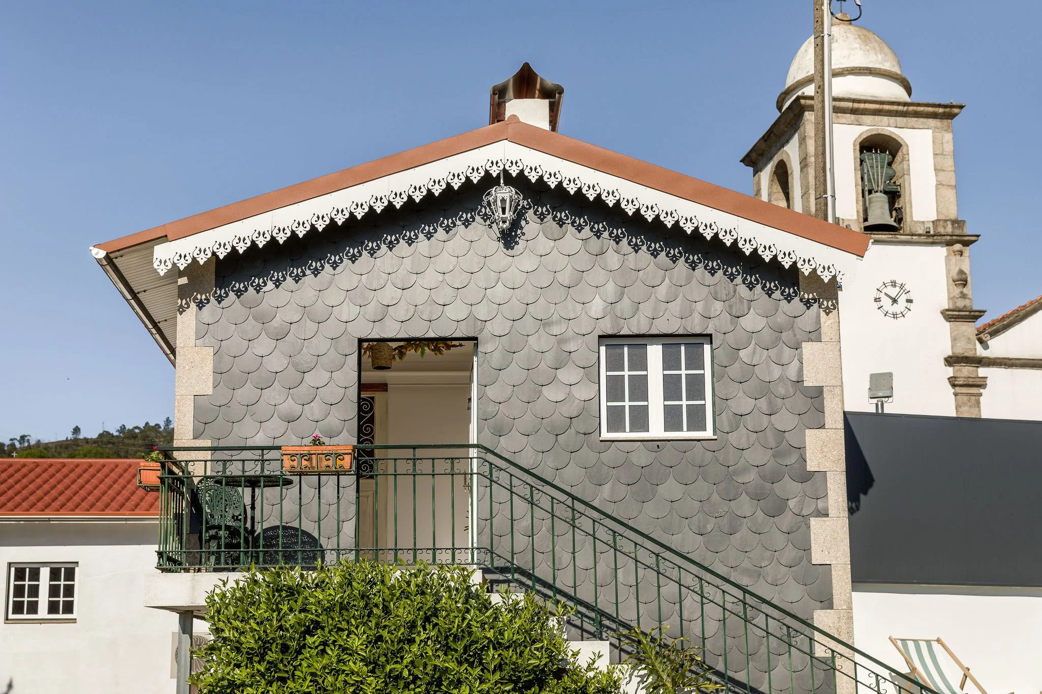 Fachada de uma casa com telhado de telhas vermelhas, paredes de tinta branca, uma varanda com guarda-corpo de ferro verde, uma janela com grades na parte inferior e uma porta de entrada aberta. Na parte superior, uma parede de escama de peixe de cor cinza, com uma janela e uma iluminação decorativa. Ao lado, uma torre de uma igreja com sino e um relógio.