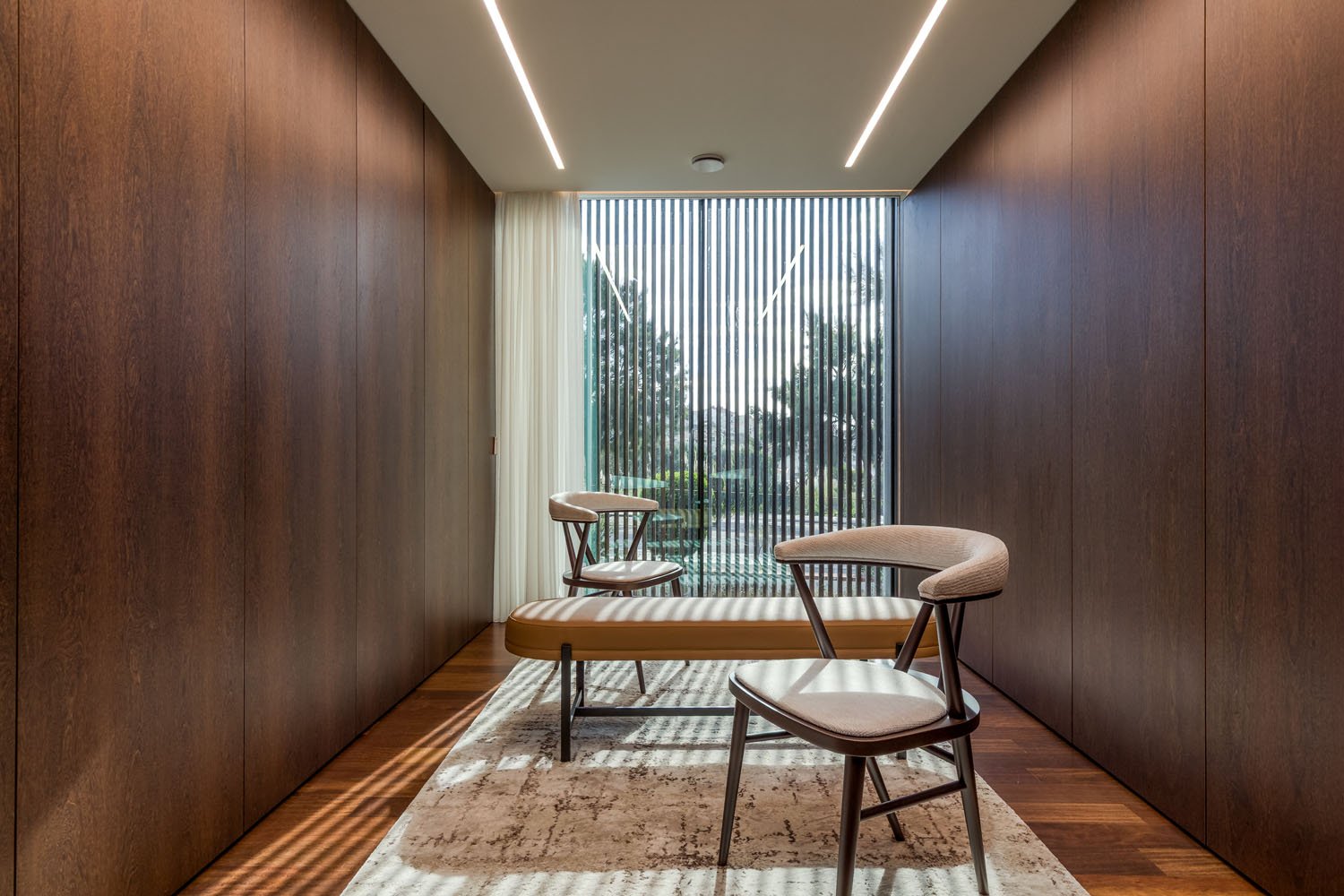 Sala de estar moderna com paredes de madeira, cortina branca, poltrona e banco de descanso com almofadas, grande janela de vidro com persianas e vista para árvores.