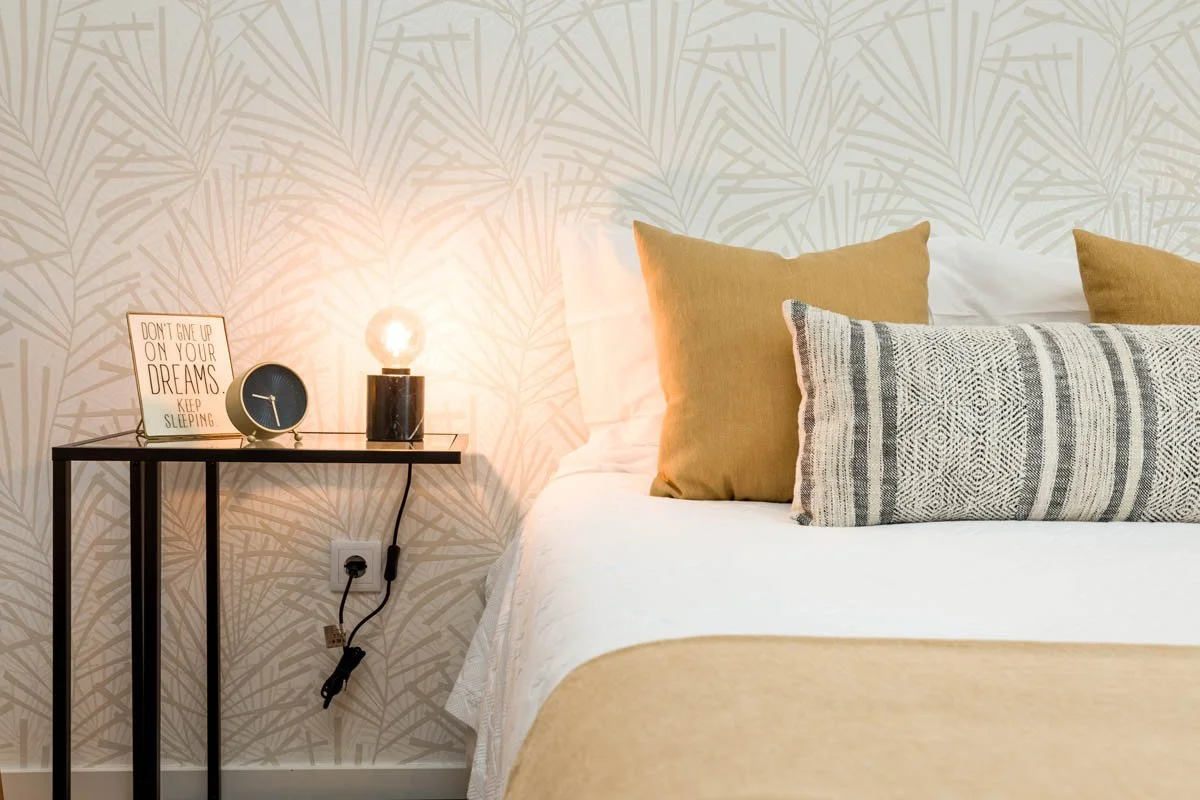Cama de casal com travesseiros bege e um travesseiro decorativo com padrão geométrico, ao lado de uma parede com papel de parede com desenho de folhas. Ao lado da cama, há uma mesa preta com um compartimento para lâmpada, um relógio preto e uma placa