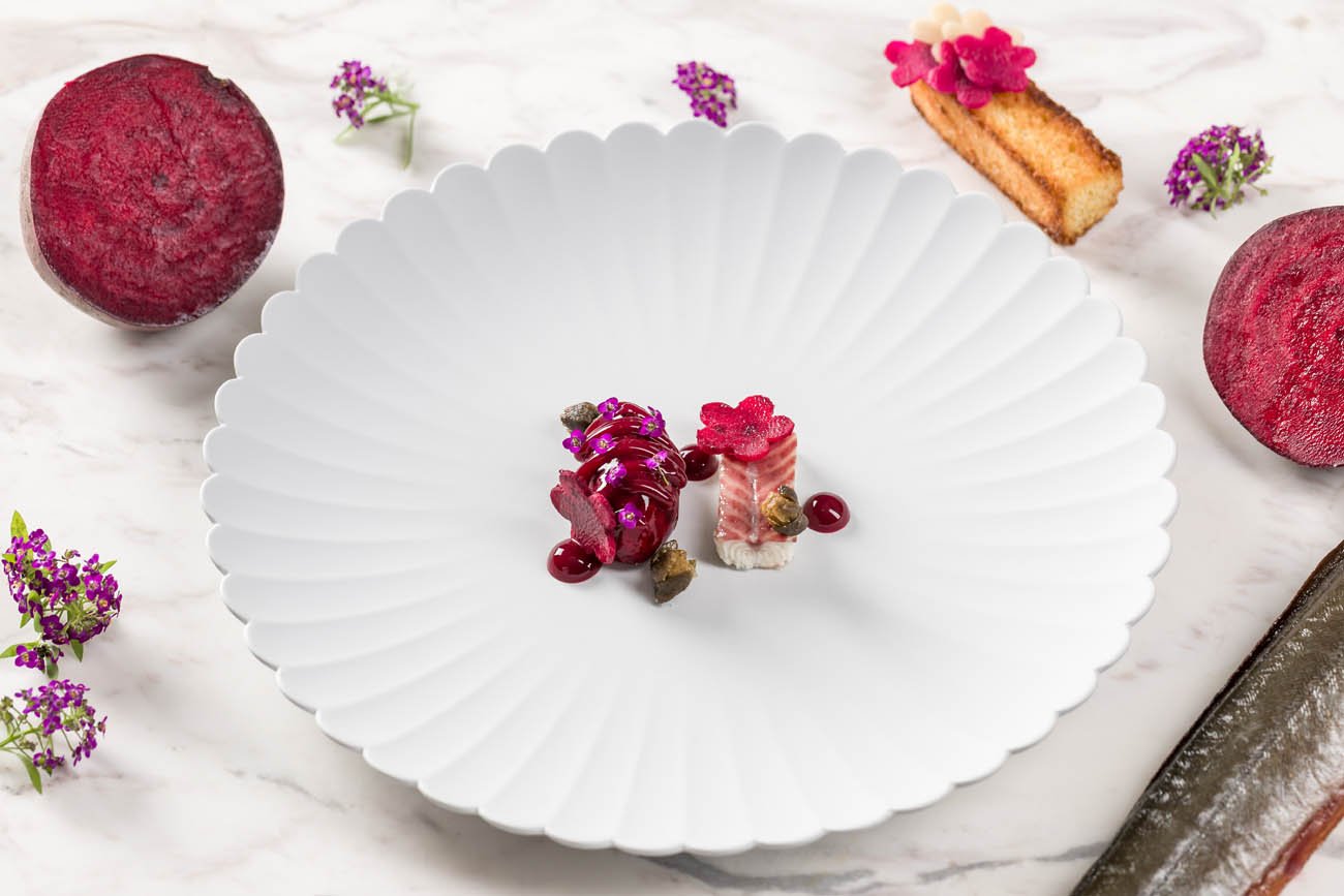 Prato com duas porções de sobremesas pequenas decoradas com flores, acompanhadas de frutas e outros doces, em uma taça branca, rodeado de frutas e flores roxas.