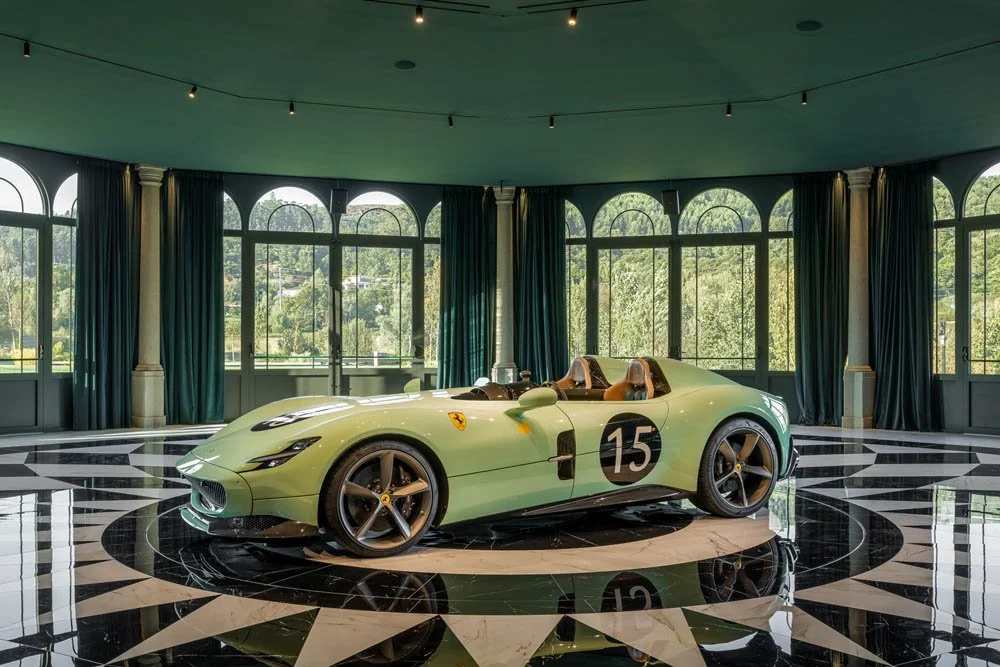 Carro esportivo verde claro com número 15, interior de couro marrom, em showroom com piso preto e branco, janelas grandes e cortinas escuras.