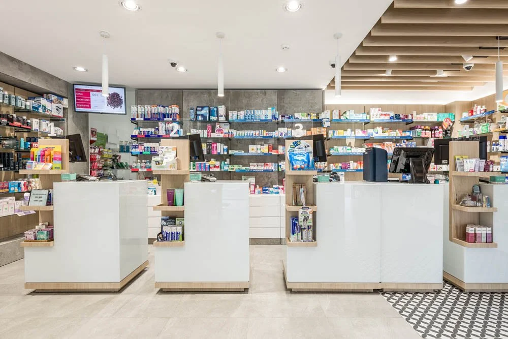Interior de uma farmácia bem iluminada com balcões brancos e prateleiras cheias de produtos de medicamentos e cosméticos.