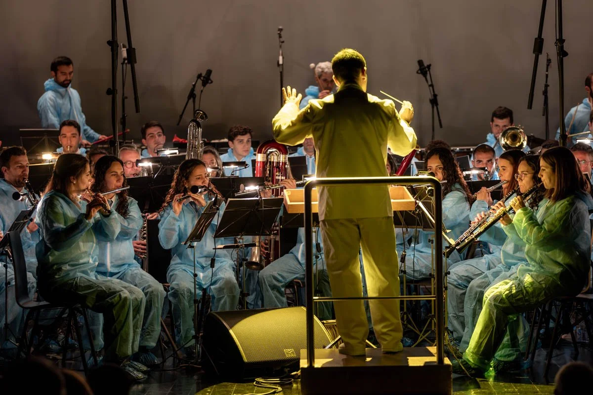 Orquestra de músicos tocando instrumentos, com destaque para o maestro na frente com uma batuta, em apresentação com iluminação de palco.