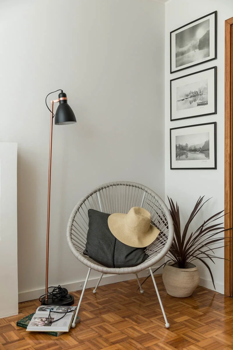 Canto de uma sala com uma cadeira de vime branco, uma planta em vaso, um abajur de pé preto com detalhes rosé, três quadros em preto e branco na parede e um chapéu de palha sobre a almofada na cadeira.