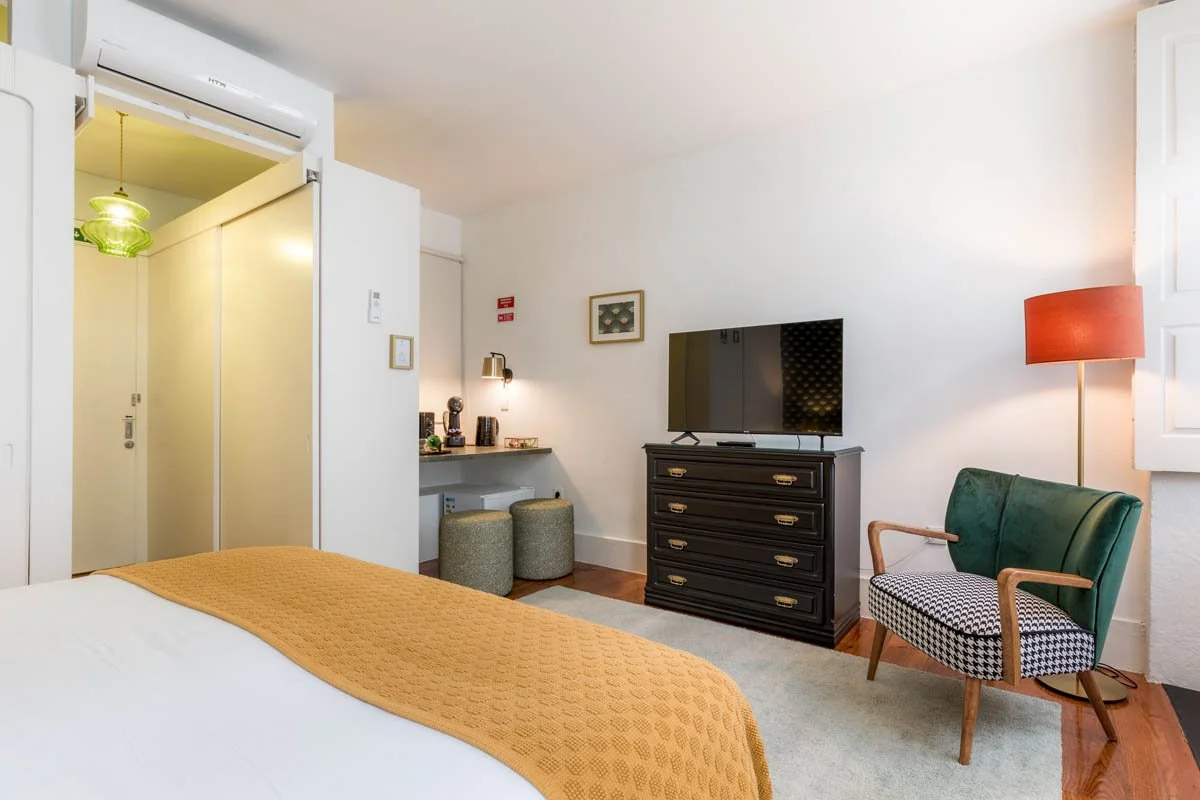 Quarto com cama, televisão em móvel de gavetas, cadeira verde, luminária laranja, bancada com duas banquetas, parede branca, iluminação suave.