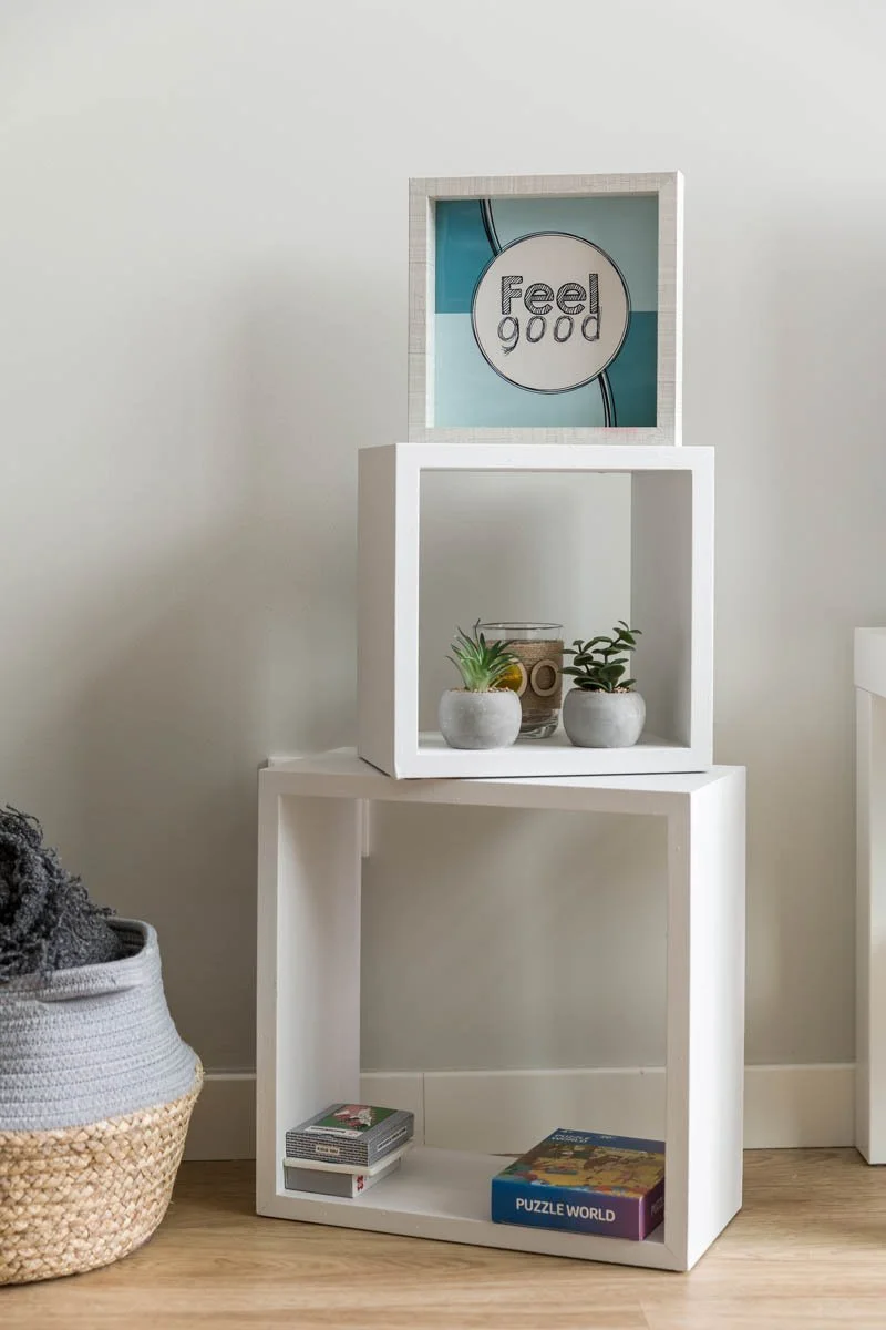 Estantes brancas em uma parede clara, decoradas com plantas em vaso, livros e um quadro com a frase 'Feel good' na parte superior.