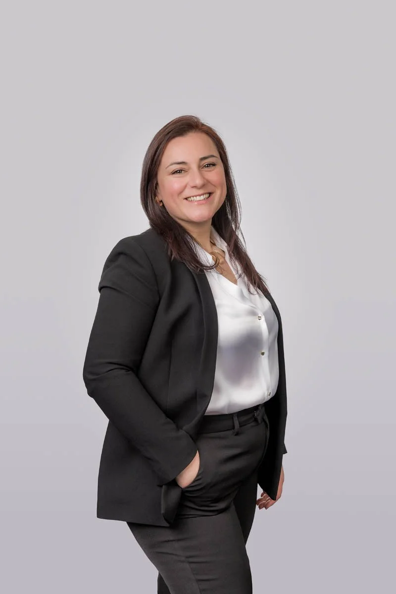 Mulher sorridente com roupa social preta e blusa branca, posando com as mãos no bolso, contra fundo cinza claro.