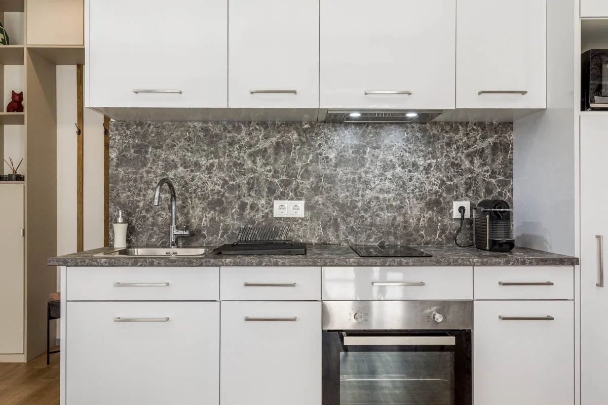 Cozinha moderna com balcão de mármore, pia, forno elétrico, máquina de café, armários brancos e parede de mármore marrom escuro.