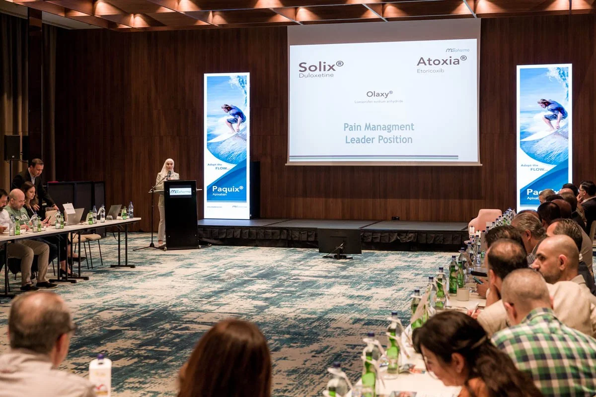 Reunião ou conferência com várias pessoas sentadas ao redor de uma mesa, uma pessoa falando no palco com um microfone, e uma tela grande exibindo o título "Pain Management Leader Position" com logos de produtos farmacêuticos.