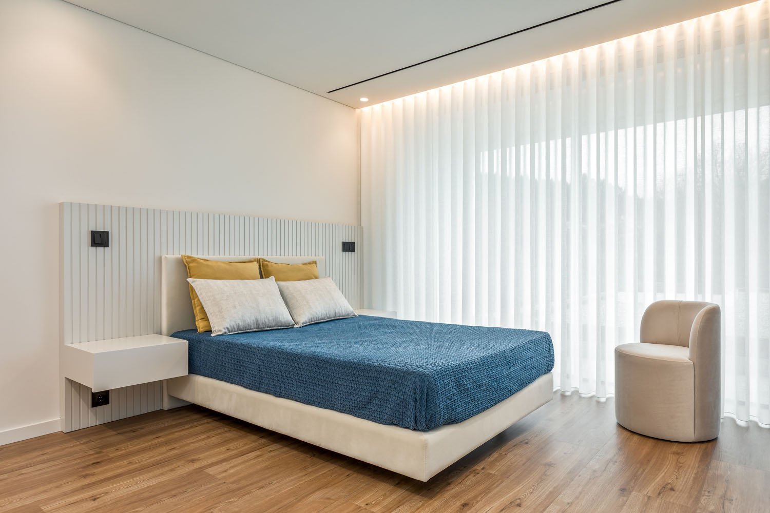 Quarto minimalista com cama de roupa de cama azul, cabeceira branca com painéis verticais, duas almofadas cinzas, duas almofadas amarelas, cadeira redonda bege, cortinas de tecido branco e piso de madeira