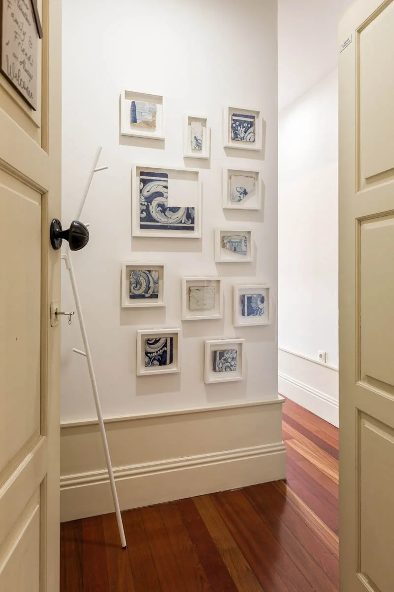 Parede com varias molduras brancas ornamentais contendo imagens de arte azul e branca, decorando uma parede interna de casa, com piso de madeira e porta aberta.