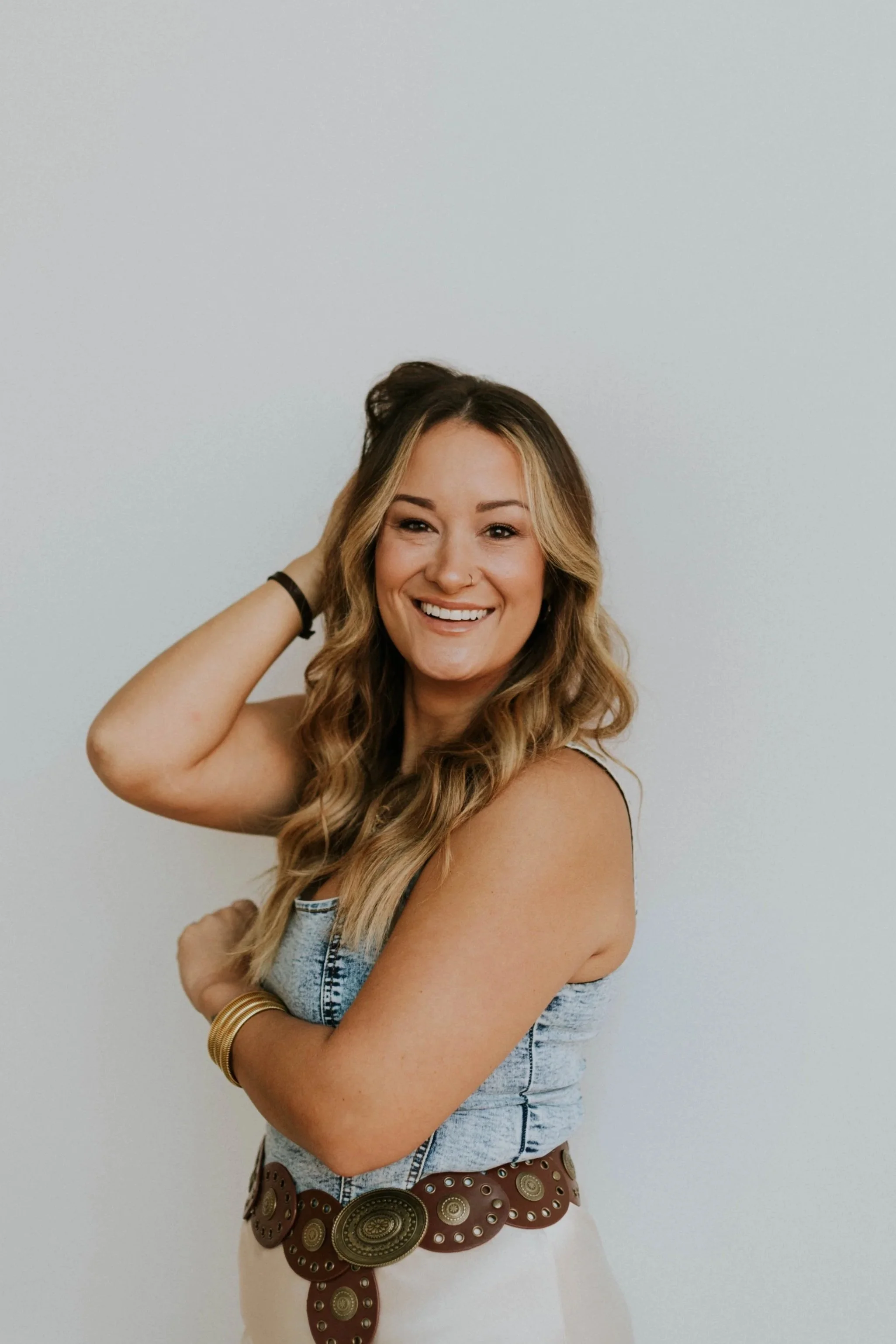 A woman with long wavy hair smiling, wearing a denim top, a large brown belt with circular metallic details, and gold bangles, posing against a plain light background.