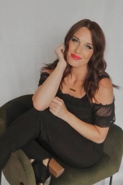 A woman with long wavy brown hair, wearing a black off-the-shoulder dress, sitting on a green armchair against a plain gray wall, smiling with her head tilted slightly and her hand resting near her face.