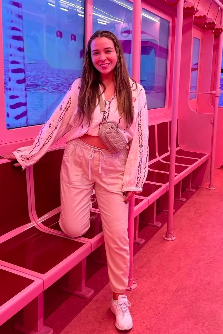 A young woman with long, wavy brown hair smiling at the camera. She is wearing a light pink crop top, matching sweatpants, and white sneakers. She has a small, gray quilted bag hanging around her neck. The background is filled with vibrant pink lighting and decorative elements, including a large digital screen and colorful hanging decorations.