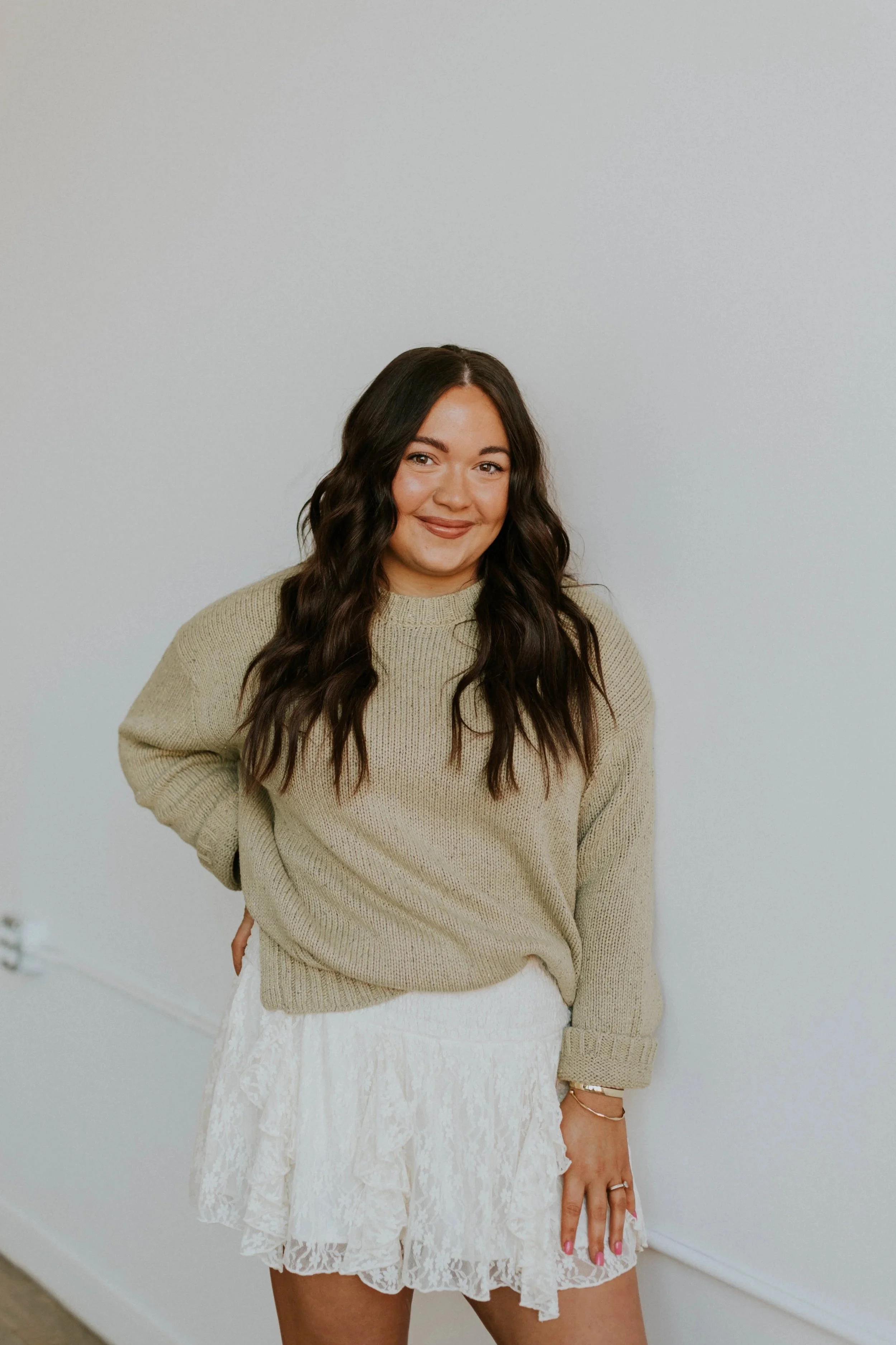 A woman with long dark hair wearing a beige sweater and a white lace skirt standing against a plain white wall, smiling at the camera.